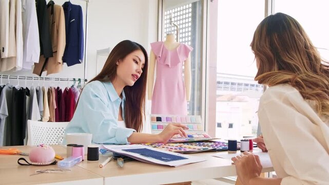 Two designers collaborate in a fashion studio, reviewing color swatches and sketches beside sewing tools and a mannequin wearing a pink dress