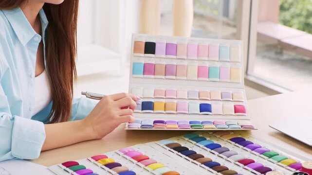 A person compares colorful fabric swatches in a sample book at a desk, selecting hues for design work beside a laptop and pen.