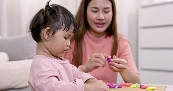 Asian single mother and young daughter playing with modeling clay together seated on floor on rolling technique using fingers during engaging early education activity at home in casual 