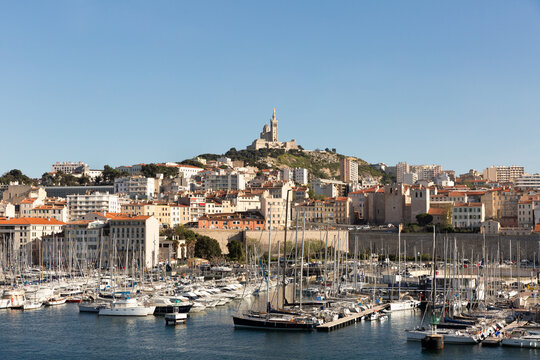 Ville de Marseille, vue sur le Vieux Port avec des bateaux &agrave; quai en premier plan et la basilique Notre Dame de la Garde sur la colline en arri&egrave;re plan, b&acirc;timents anciens et grand ciel bleu