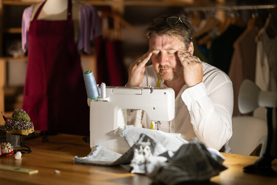 Overworked middle-aged male tailor sitting on table next to sewing machine