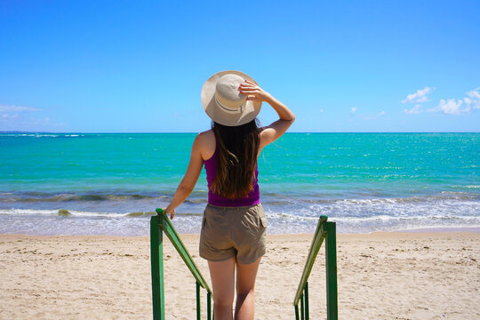Paradise beach concept. Back view of traveler woman goes down the stairs towards crystalline water of the beach.