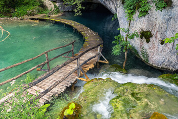 Naklejka premium Wooden boardwalk curves turquoise lake enter cave rocky cliff covered moss greenery. Clear stream flows stones form soft cascade serene untouched national park landscape tranquil natural atmosphere