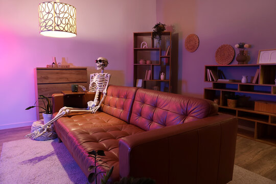 Human skeleton model with sunglasses and cup sitting on couch in living room at night