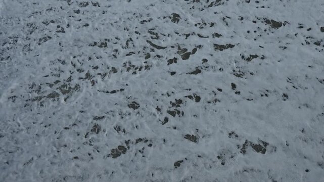 Foam washing over black pebble beach, closeup texture study revealing frothy lace, wet basalt stones, minimal moody palette