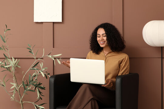 Young African-American woman using laptop in armchair near brown wall at home