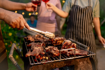 Hands grilling steaks on barbecue with tongs at outdoor garden party  people toast red wine in background, warm evening lights, casual summer gathering. © Thitaporn