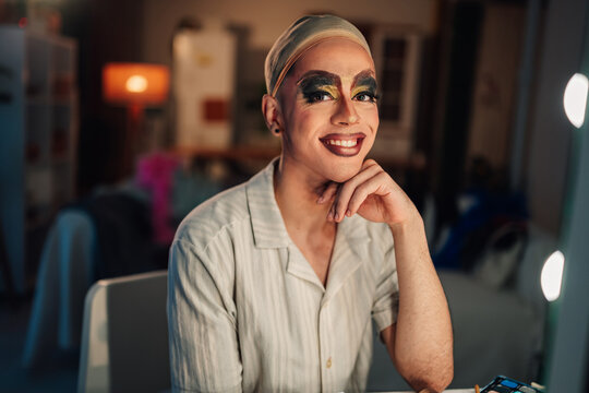 Drag performer smiling by vanity mirror, confidently preparing for performance with bold makeup and wig cap