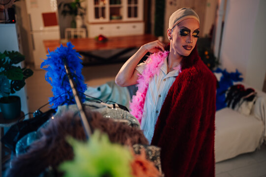 Drag queen in makeup and wig cap getting ready, wearing a pink boa and red coat amidst colorful feather boas