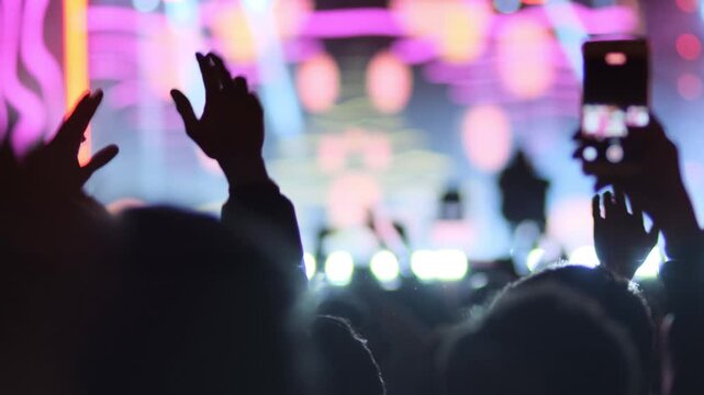 Hands of dancing people raised up. Many people dance to fast rhythmic electronic music accompanied by bright light effects. Crowd cheering and jumping on dance floor in front of stage with DJ.