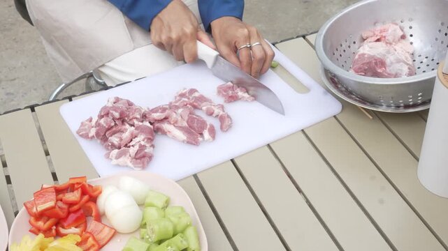 Hands cutting raw pork on cutting board beside bowl of chopped vegetables and colander of meat on outdoor table, food prep for cooking.