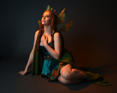 portrait of female model wearing fantasy fairy costume, green dress, cloak, butterfly wings and crown. full length sitting pose, kneeling on floor, dark studio background.