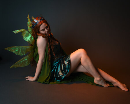 portrait of female model wearing fantasy fairy costume, green dress, cloak, butterfly wings and crown. full length sitting pose, kneeling on floor, dark studio background.