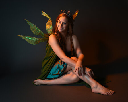 portrait of female model wearing fantasy fairy costume, green dress, cloak, butterfly wings and crown. full length sitting pose, kneeling on floor, dark studio background.