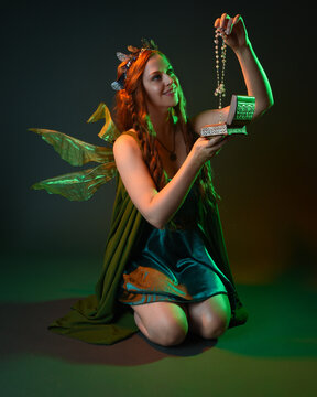 portrait of female model wearing fantasy fairy costume, green dress, cloak, butterfly wings and crown. Full length kneeling pose holding secret jewellery box, isolated on dark studio background