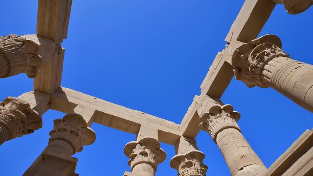 Upward view of ornate floral capitals on Ptolemaic columns at Philae Temple, Aswan Egypt. Elegant ancient architecture against vivid blue sky. Travel and history content.