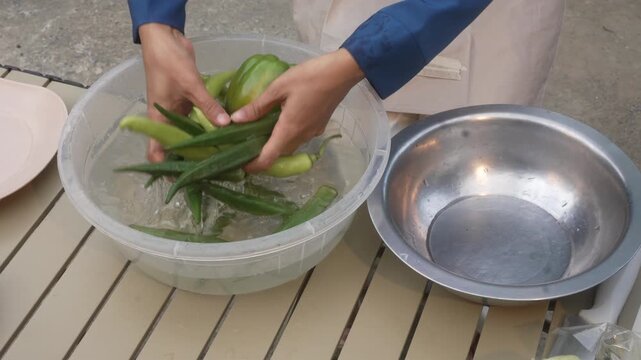 Hands washing fresh okra and green peppers in a plastic tub of water on an outdoor table, with a metal bowl beside, food prep and hygiene concept.
