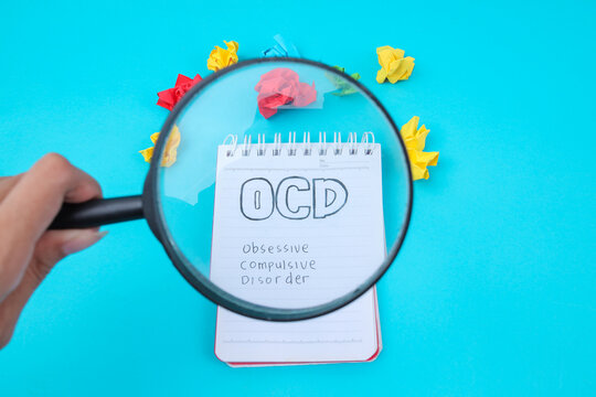 Hand Holding Magnifying Glass Over Notepad With OCD Text And Crumpled Papers For Mental Health Concept