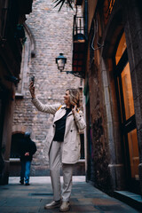 Obraz premium Full length smiling woman taking photo with mobile phone in city alley. Mature female traveler exploring urban landmarks, digital nomad lifestyle, authentic representation, copy space