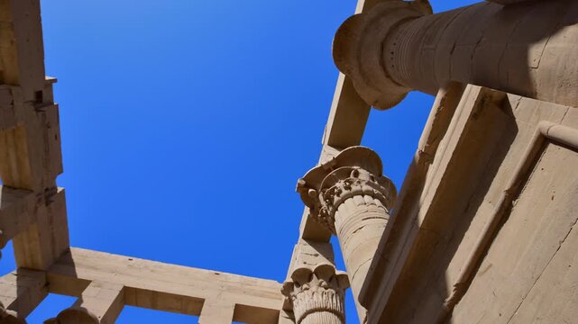 Dramatic upward view of ornate papyrus columns with carved floral capitals and stone lintels at Philae temple, Aswan, Egypt, against vivid deep blue sky.