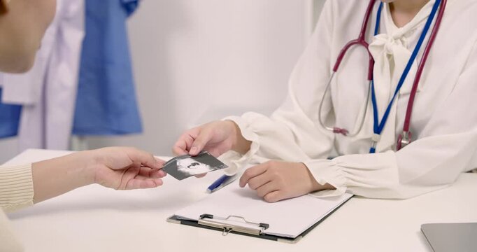 Obstetrician receiving ultrasound image from Asian pregnant woman during maternity consultation in clinic showing happy interaction medical support and positive relationship in third trimester