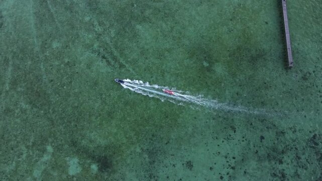 Aerial drone shot of a speedboat towing a banana boat over clear green tropical waters, with a small pier visible in the frame. Cinematic top view capturing summer vacation, water sports activity, and