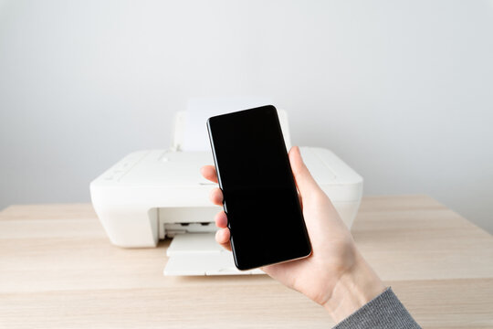 Person holds a smartphone in front of a printer on a wooden desk, wireless mobile printing and connectivity for a modern home office, highlighting convenience, productivity and simple workflow.