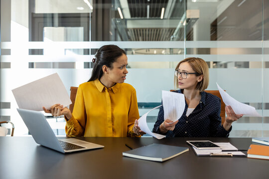 Two stressed businesswomen at a modern office table, one gesturing emotionally with documents while the other listens worriedly, illustrating workplace conflict and communication breakdown