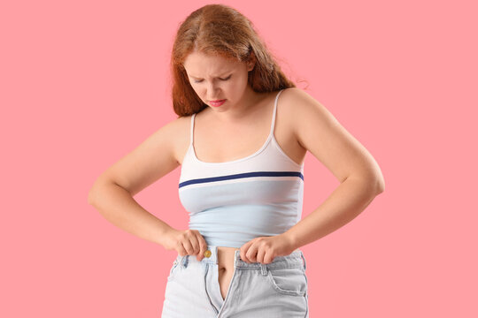 Young woman trying to button tight shorts on pink background. Weight gain concept