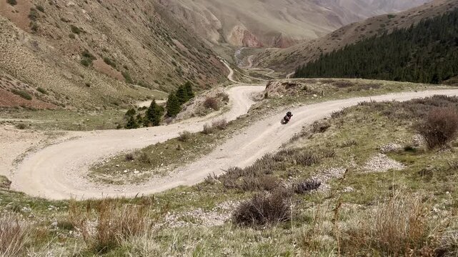 A motorcycle traveler navigates the famous winding switchbacks leading to Song-Kul Lake in Kyrgyzstan. The gravel road snakes through a vast mountain valley under a dramatic alpine landscape