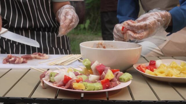 Hands preparing meat and vegetable skewers on a table outdoors, wearing gloves, with bowls and plates of chopped ingredients for a barbecue or picnic.