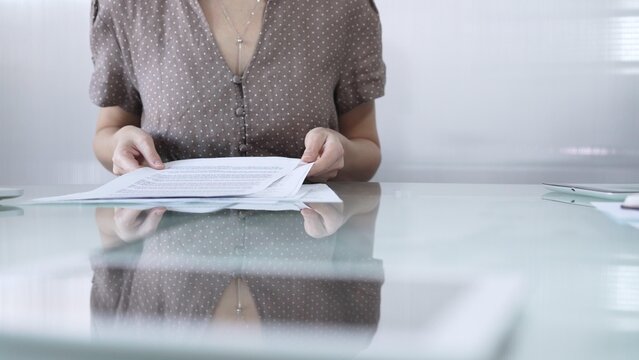 Businesswoman or accountant in brawn dotted blouse is going through paperwork while holding documents at a glass desk in a well lit, contemporary office setting. Business people concept
