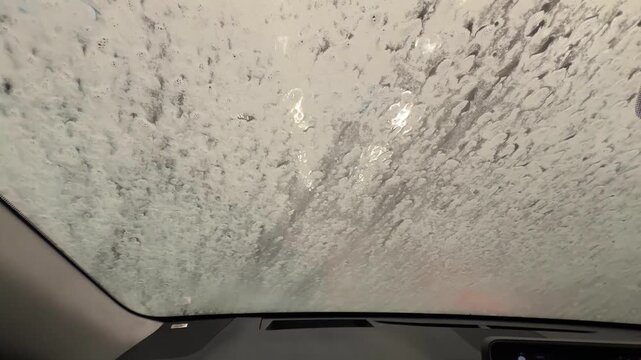 Foam and water drops flowing over car windshield during automatic car wash. View from inside vehicle through wet glass with bubbles and blurred lights outside.