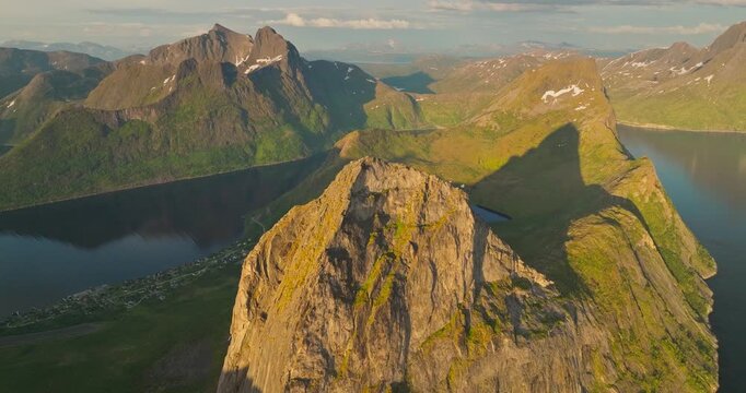 Sheer granite cliffs rise from the Arctic waters in Northern Norway, Senja island, breathtaking drone view of Segla mountain range during midnight sun with snow capped peaks reflecting in calm water