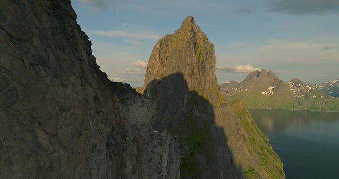 Person stands on rock facing the iconic Segla mountain peak on Senja Island, Norway at Hesten hike summit. Drone flies over revealing the sheer cliffs rising from the water