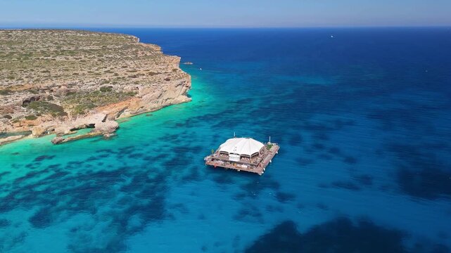 Noma Island, anchored off the coast of Comino in Malta&rsquo;s Mediterranean archipelago, features a floating venue with outdoor lounge areas, bar and pool in clear turquoise water in an aerial view.
