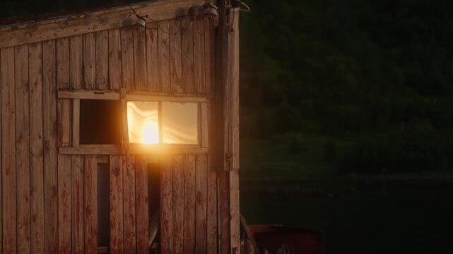 Warm golden sunlight filters through and reflects on the window of an old wooden shed, creating a cinematic play of light, texture, and rustic atmosphere in a serene rural setting.