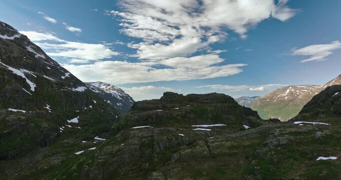 Drone rising from behind a granite mountain, revealing turquoise Oldevatnet Fjord waters surrounded by snow-capped peaks under a clear blue summer sky in Norway.