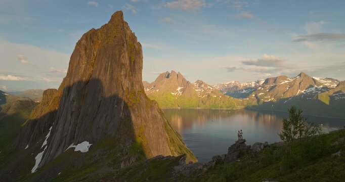 People standing on rock in front of of majestic Segla mountain peak with panoramic views of the fjord waters reflecting surrounding snowcapped mountains on Senja Island Norway during midnight sun