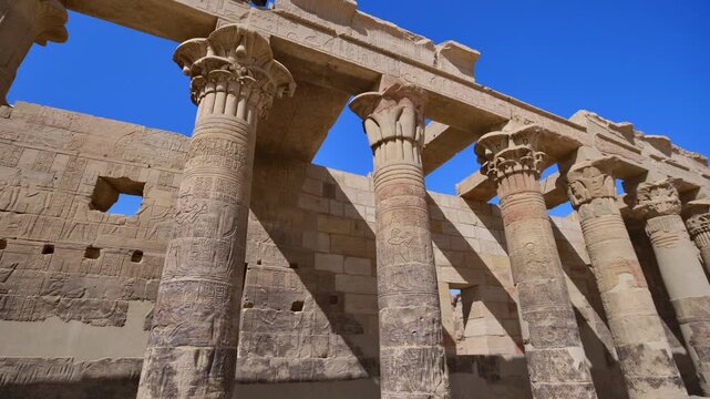 Dramatic upward view of ornate papyrus columns with carved floral capitals and stone lintels at Philae temple, Aswan, Egypt, against vivid deep blue sky.