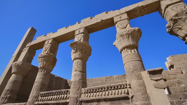 Looking up through carved sandstone columns with decorative papyrus capitals and stone crossbeams at Philae temple, Aswan, Egypt, clear blue sky above.