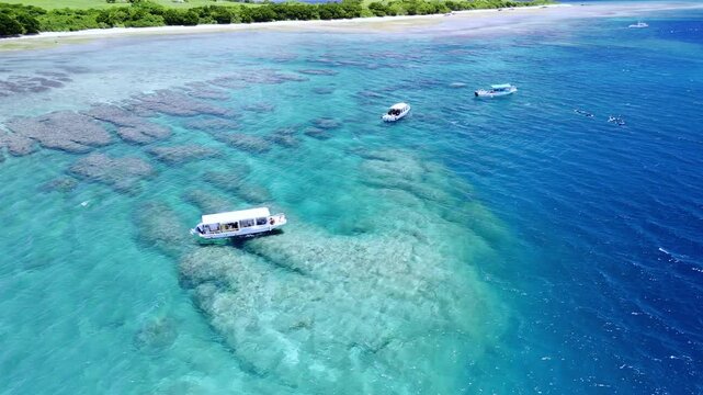 Drone shot showcasing glass-bottom boats and snorkelers exploring vibrant coral reef structures in the transparent, multi-toned blue waters of Okinawa, Japan.