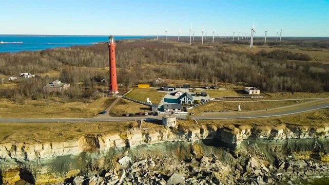 Beautiful aerial perspective of the historic Pakri lighthouse standing atop a rugged cliff on the Paldiski peninsula, with a modern wind farm visible along the coastline under a clear blue sky