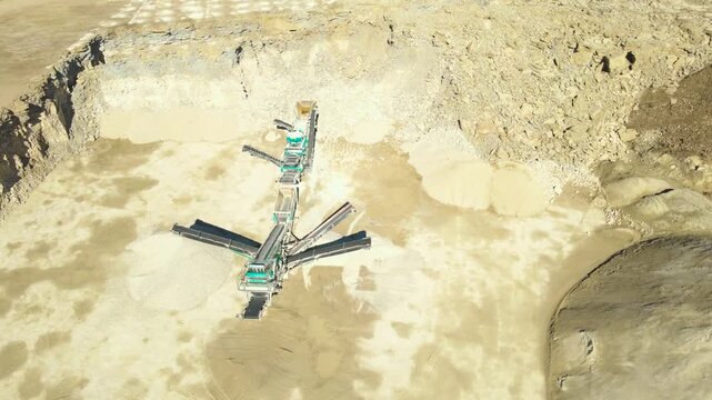 Aerial drone shot slowly rotating above an open pit limestone quarry, showing heavy machinery such as a stone crusher, conveyor belts, and piles of sand and gravel on a sunny day