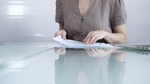 Businesswoman or accountant in brawn dotted blouse is going through paperwork while holding documents at a glass desk in a well lit, contemporary office setting. Business people concept