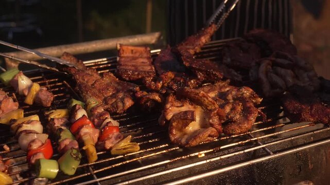Close-up of assorted meats and vegetable skewers grilling on a barbecue at night, sizzling on metal grate with tongs and warm firelight, outdoor cookout scene.