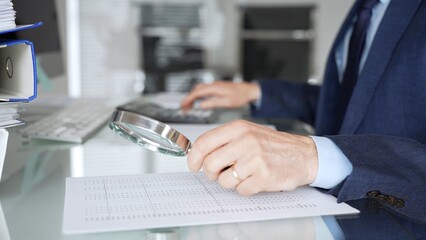 Fototapeta premium Hand holding magnifying glass, inspecting financial document, performing audit and business analysis. Audit and taxes in business