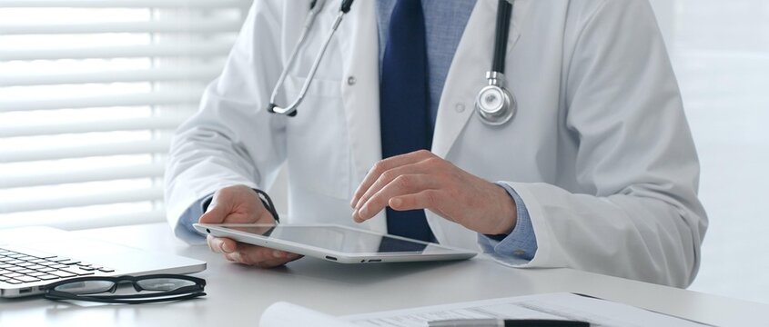 Doctor wearing lab coat and stethoscope using digital tablet while sitting at desk in medical office with laptop, eyeglasses, and medical chart nearby. Medicine and health care concept