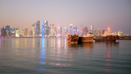 Naklejka premium Traditional dhow boats illuminated at night on the calm waters of Doha Corniche, with the vibrant West Bay skyline glowing in the background and colorful reflections shimmering across the Arabian Gulf