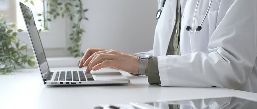 Medical professional wearing white lab coat typing on laptop, documenting patient information with clinical precision at workstation. Medicine and health care concept